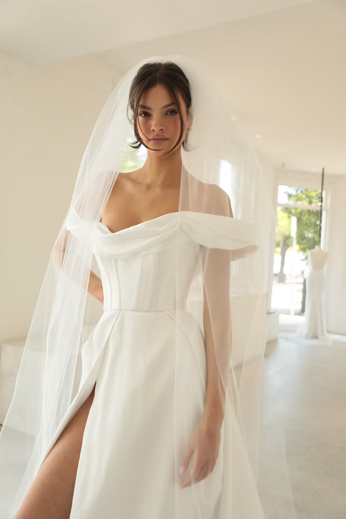 Bride in white gown with off-the-shoulder neckline and high slit, wearing a veil in a bright room.
