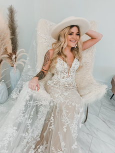 Woman in wedding dress and hat, seated in a chair, arm raised, smiling.