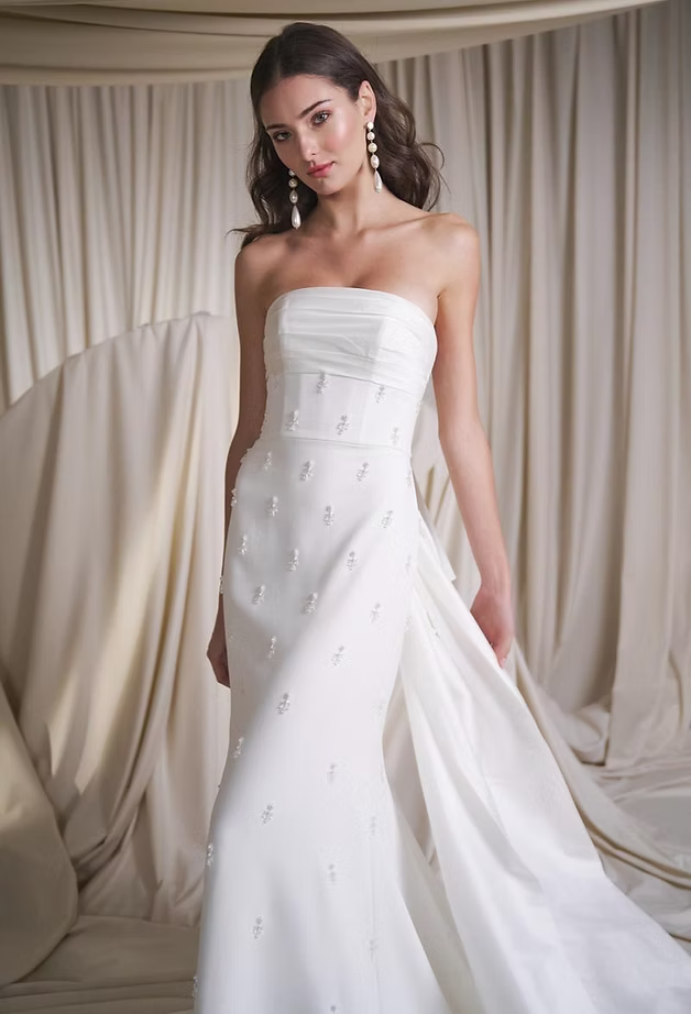 Woman in a white strapless wedding dress with pearls, standing indoors.