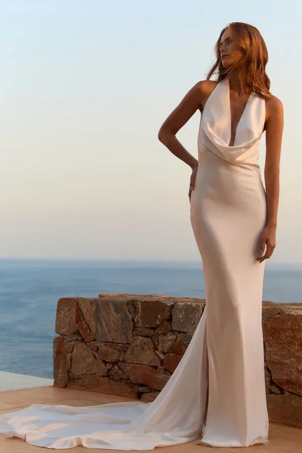 Woman in white wedding dress with cowl neckline poses outdoors, ocean view.