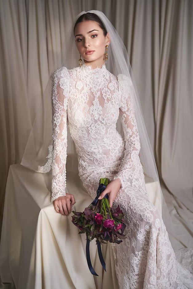 Bride in lace gown holds bouquet; beige backdrop.
