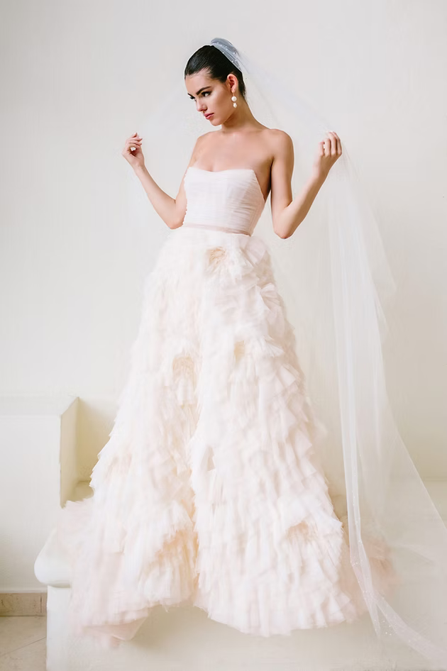 Bride in strapless gown with ruffled skirt and veil, standing on a white staircase.