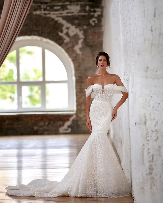 Woman in an off-the-shoulder white wedding dress, posing near a window in a rustic room.