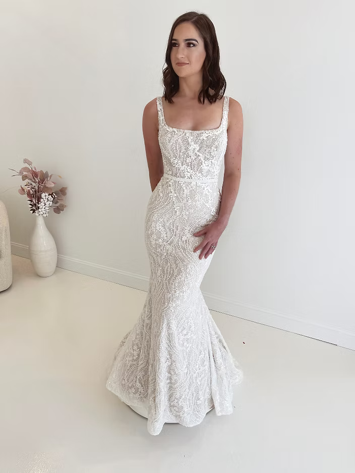 Woman in a white, lacy mermaid wedding dress, standing in a bright, white room.