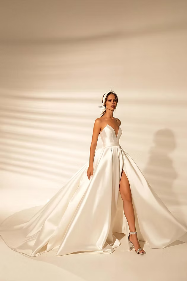 Woman in a white satin wedding gown with a high slit, standing in a studio with shadows.