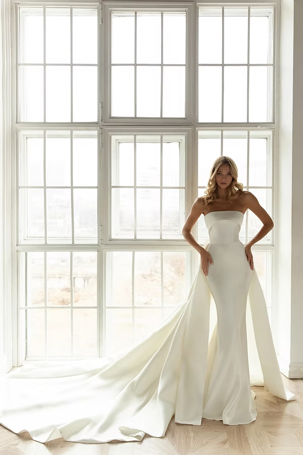 Woman in a strapless white wedding dress with a train, standing by a large window.