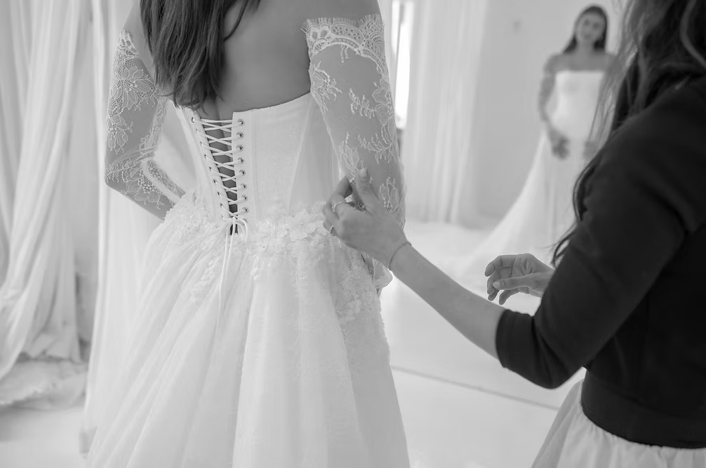 Woman in wedding dress having it adjusted; mirror reflection; lace sleeves, corset back.
