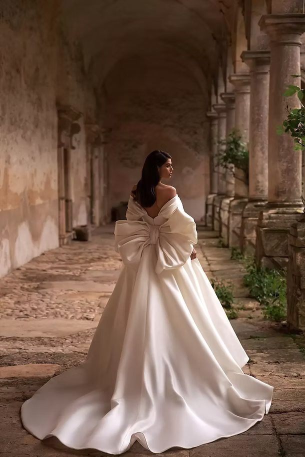 Bride in white satin gown with large bow, walking in arched stone hallway.