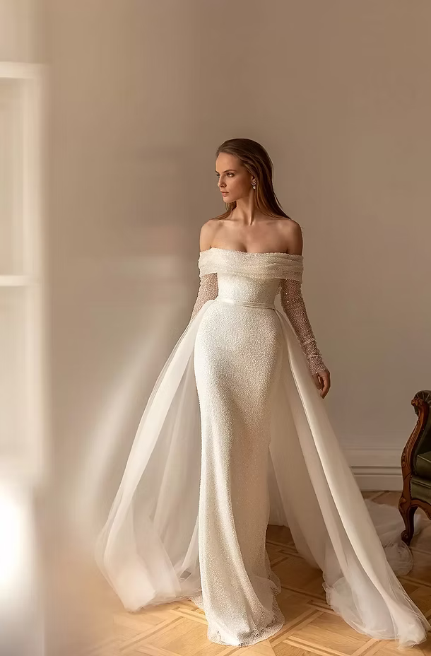 Bride in off-the-shoulder gown with long sleeves and train, standing near a window.