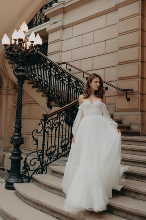 Woman in white gown descends grand staircase, holding the skirt. Ornate black lamppost, architectural background.