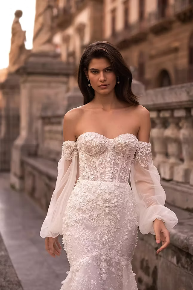 Woman in a white, off-the-shoulder wedding dress, walking outdoors in a stone architectural setting.