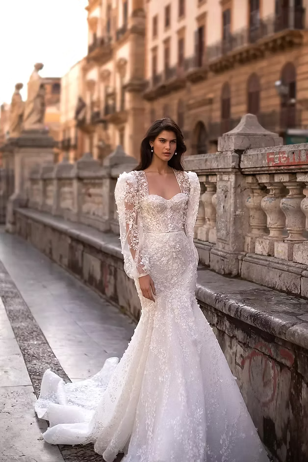 Woman in a white, beaded wedding gown with a long train stands on a stone walkway.