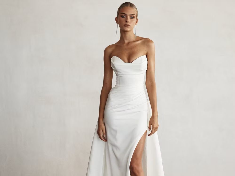 Woman in strapless white wedding dress with slit and train, standing against a white background.