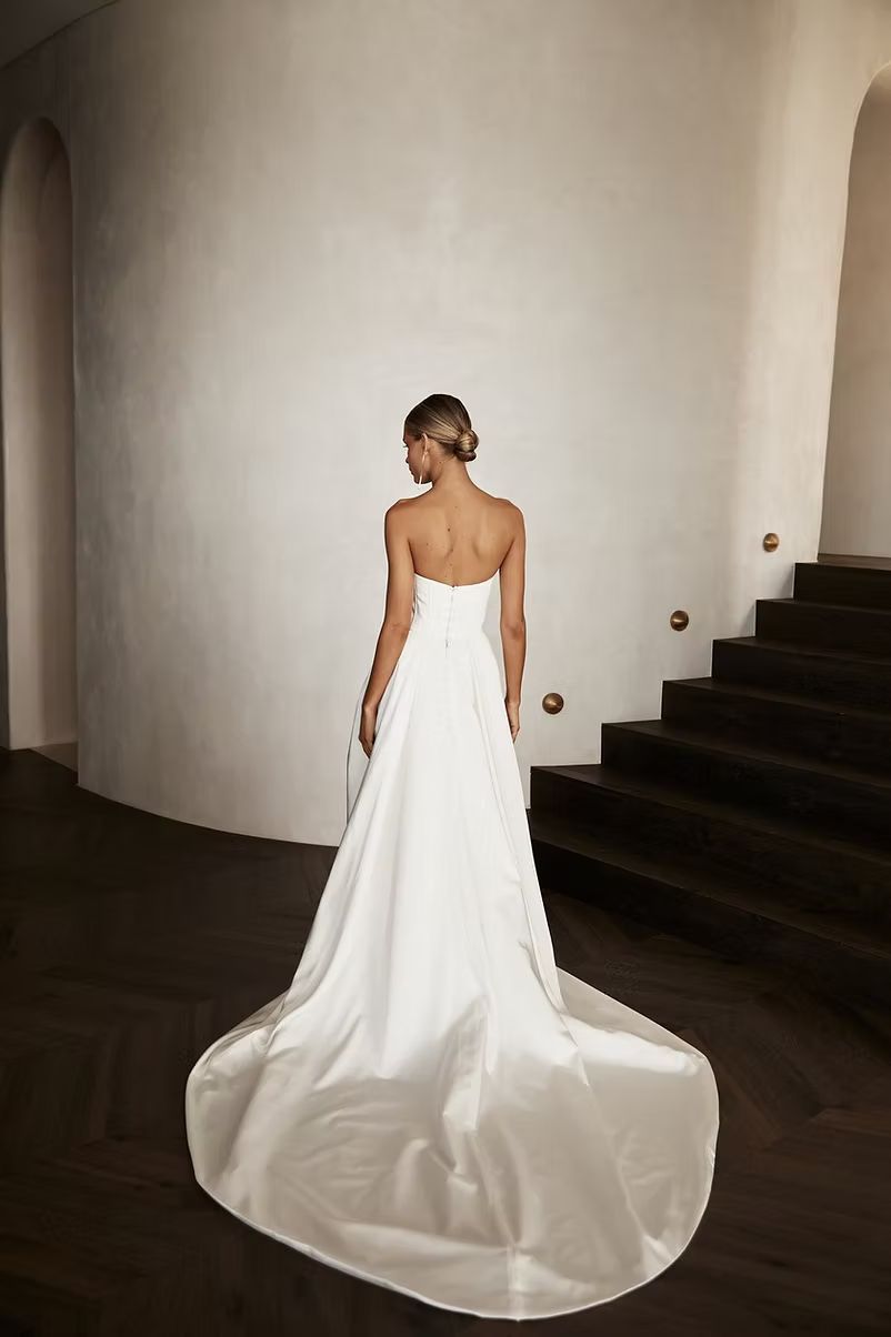Bride in strapless white gown stands near staircase, back to the viewer.