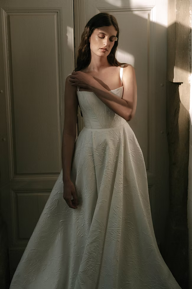 Woman in white wedding dress, standing near a door, eyes closed, sunlit.