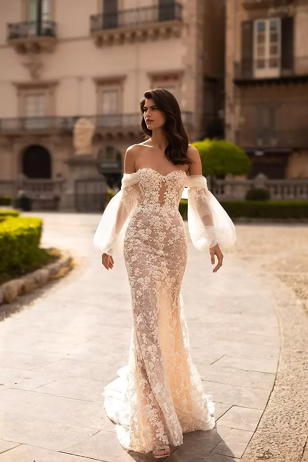 Woman in a lace off-the-shoulder wedding dress, standing outside in front of a building.