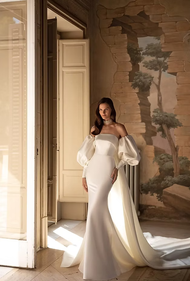 Woman in white off-the-shoulder gown with long train poses in front of painted wall, sunlight streams in.