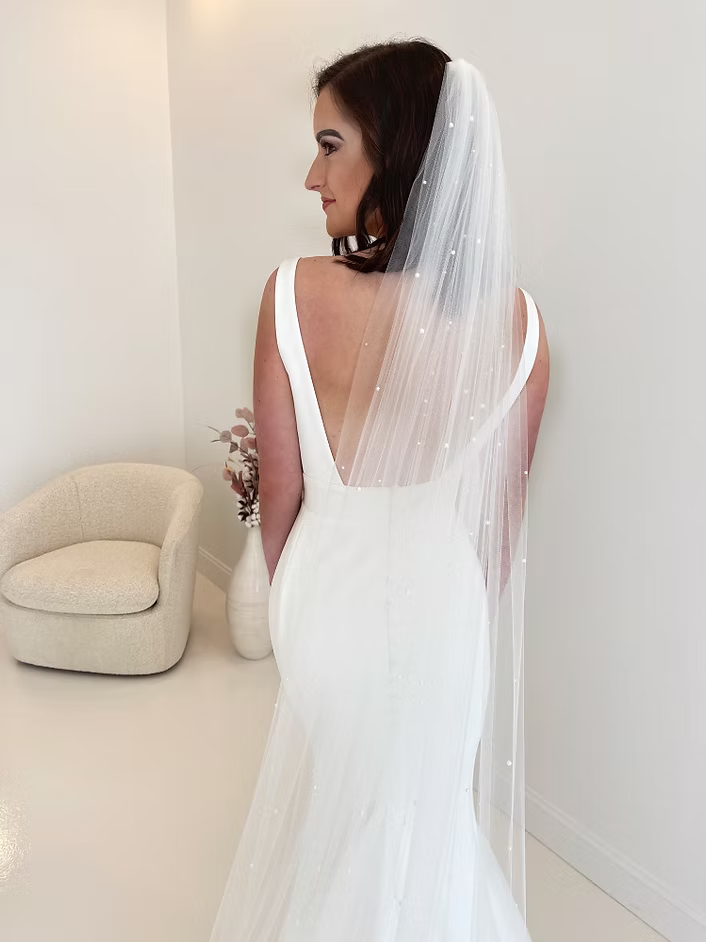Bride in a white dress with a pearl-studded veil, standing indoors, turned to the side.