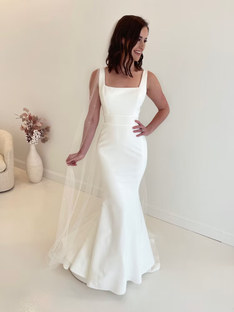 Woman in white wedding dress with square neckline, veil, smiles.