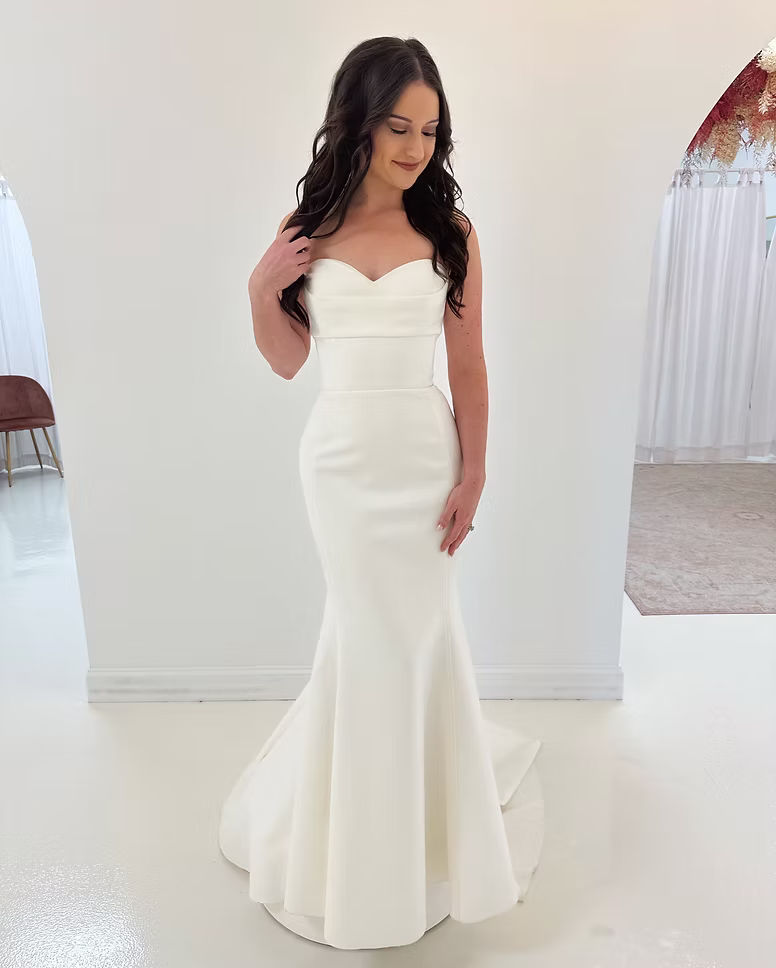 Woman in a white, fitted mermaid wedding dress with sweetheart neckline. She smiles, in a shop setting.