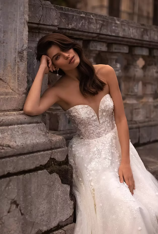 Woman in strapless wedding dress leans against a stone wall, looking at the camera.