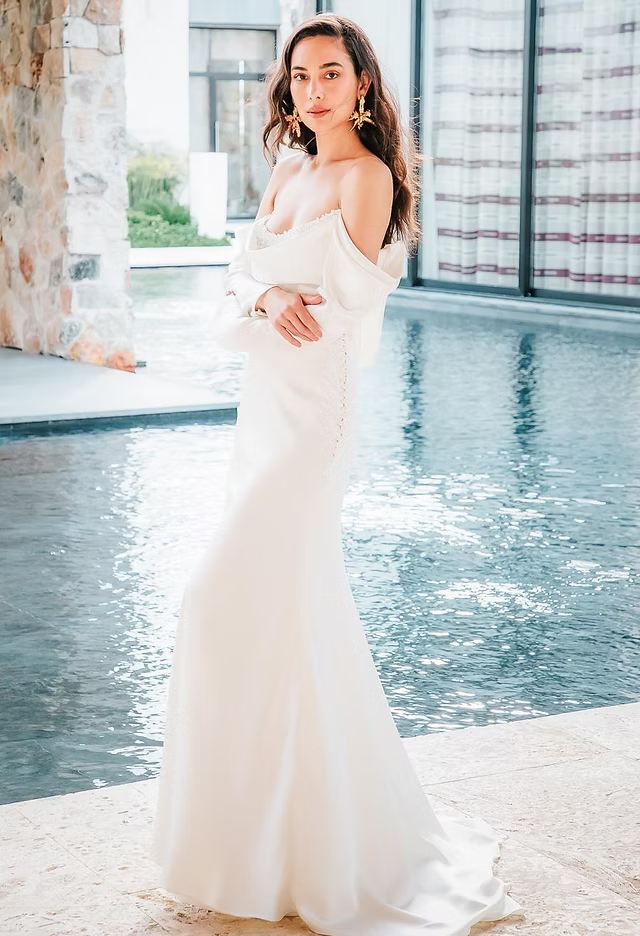 Woman in white off-the-shoulder dress stands by water. Leaning in, wearing gold earrings.