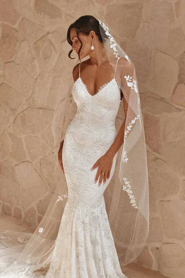 Bride in white lace mermaid gown, veil with floral embroidery, standing before stone wall.