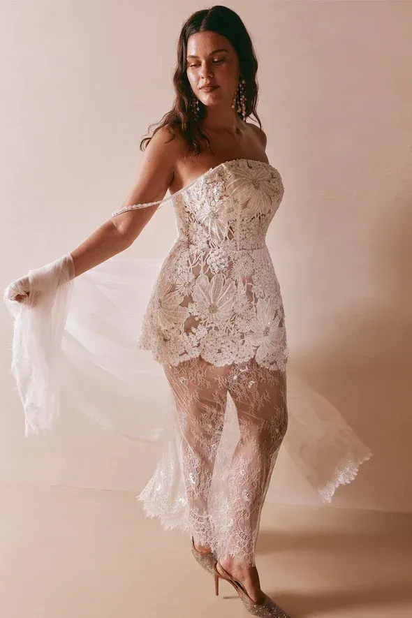 Woman in white lace bridal outfit, smiling, arms outstretched, studio shot.