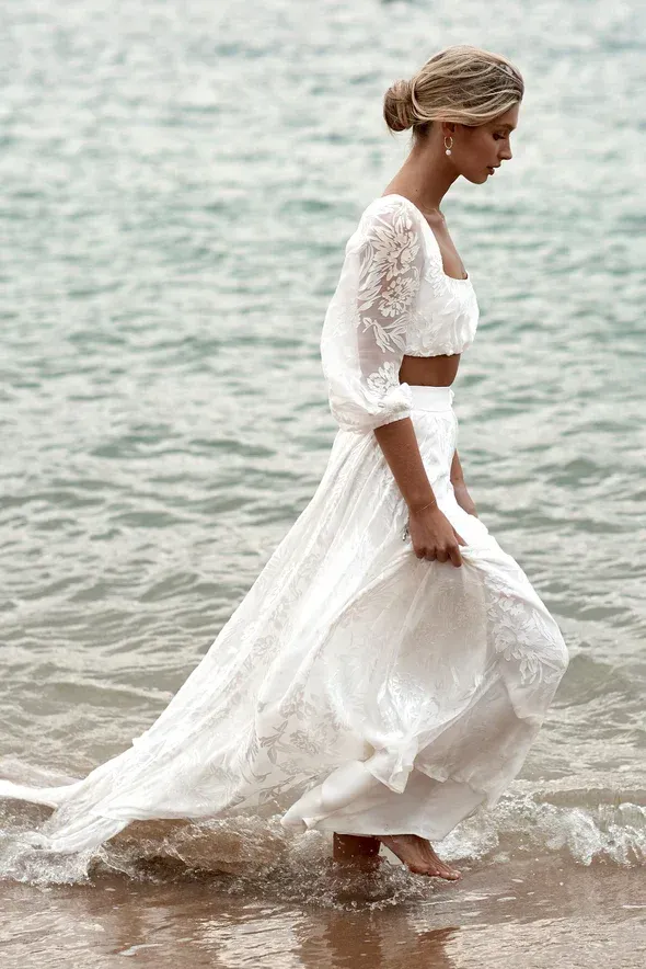 Woman in white lace outfit, barefoot, walking in ocean.