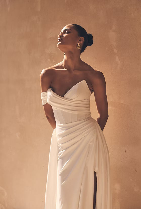 Woman in white off-shoulder dress, eyes closed, arms behind back, sunlit against beige wall.