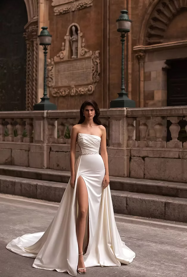 Woman in strapless white gown with high slit, poses on steps in front of ornate stone building.