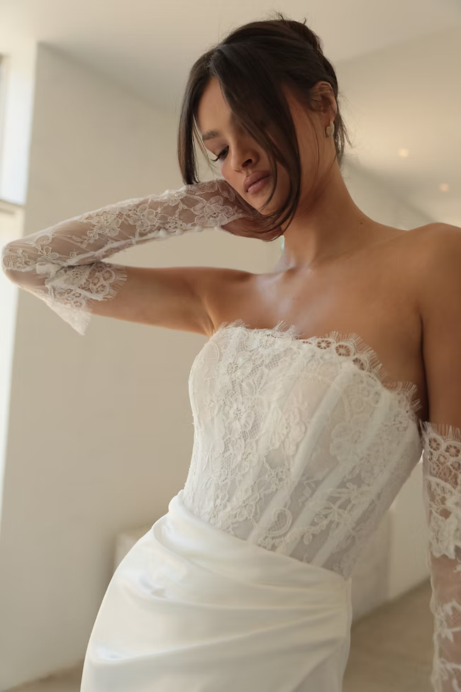 Woman in a strapless lace wedding dress with lace gloves, soft expression, leaning.