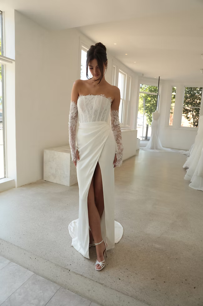 Woman in a white wedding dress with lace gloves and high slit. Indoors, neutral setting.