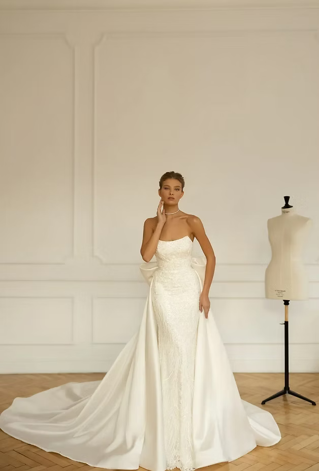 Woman in white strapless wedding dress with long train, standing by a mannequin in a light room.