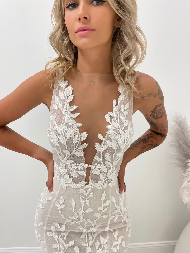 Woman in a white, floral lace wedding dress with a plunging neckline, indoor setting.