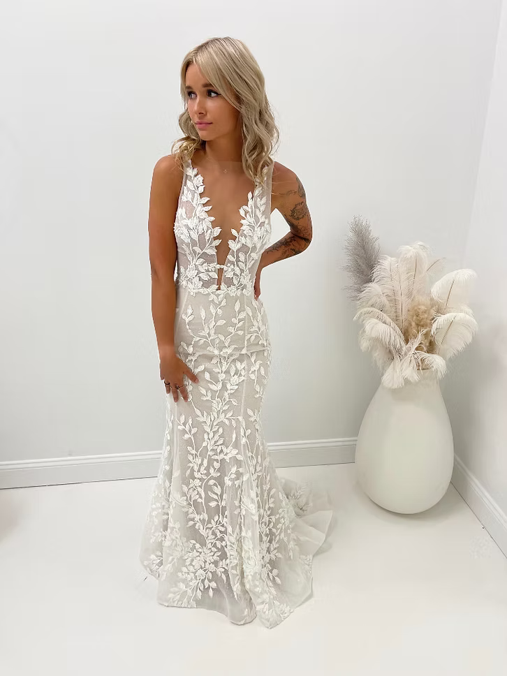 Woman in a white, floral lace wedding dress, stands indoors, near a vase with dried flowers.