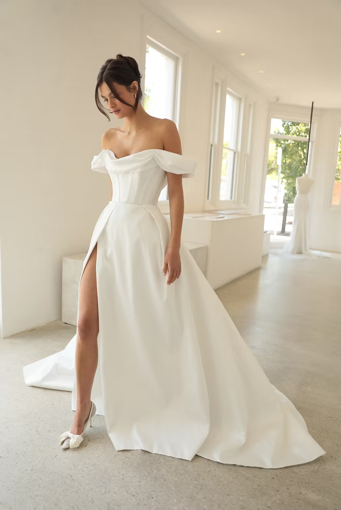 Woman in an off-shoulder white wedding dress with a thigh-high slit, posing in a bright white room.