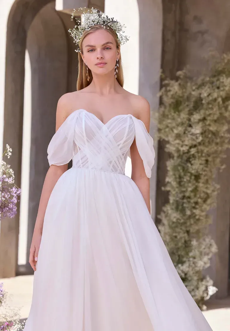 Woman in white off-the-shoulder wedding dress with floral crown, standing outdoors near archway.