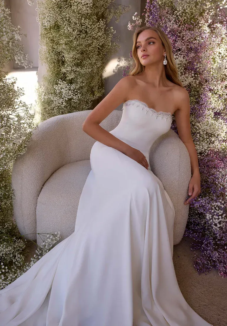 Woman in strapless white wedding dress sitting in a curved chair, surrounded by flowers.