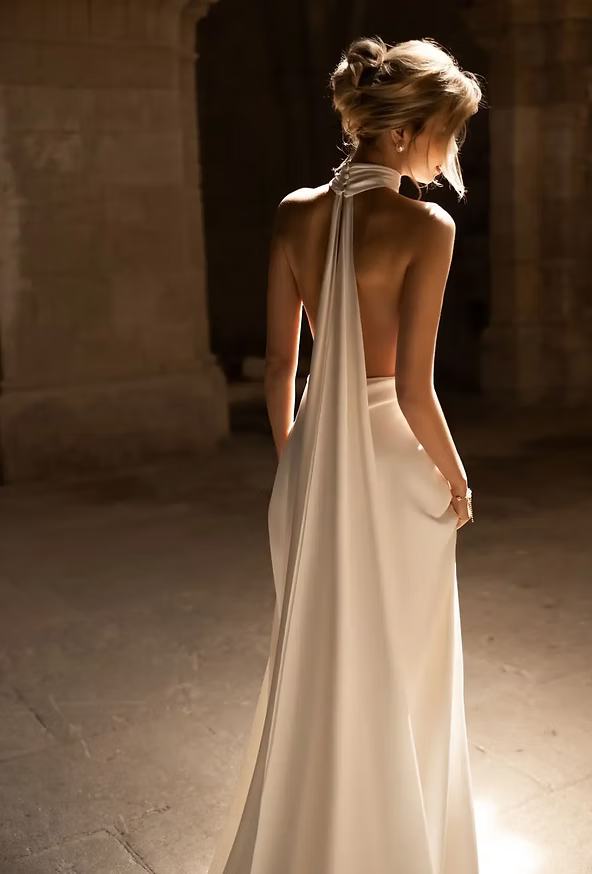 Woman in white halter gown, back to camera, standing in dimly lit stone room.