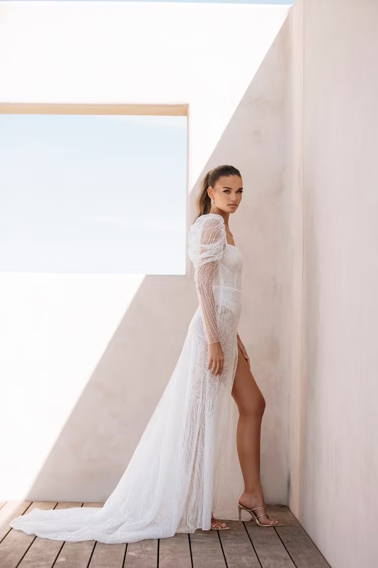 Woman in white wedding dress with thigh-high slit, standing outdoors.