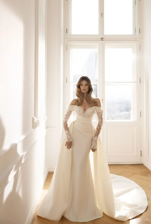 Woman in white off-shoulder wedding dress with long sleeves and train, standing by a window.