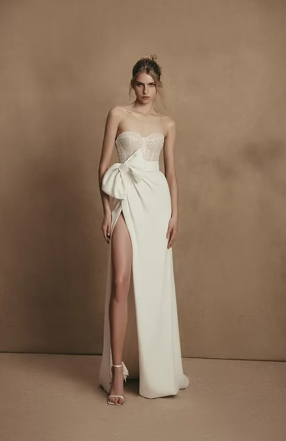 Woman in white strapless gown with thigh-high slit and oversized bow. Light hair, neutral background.
