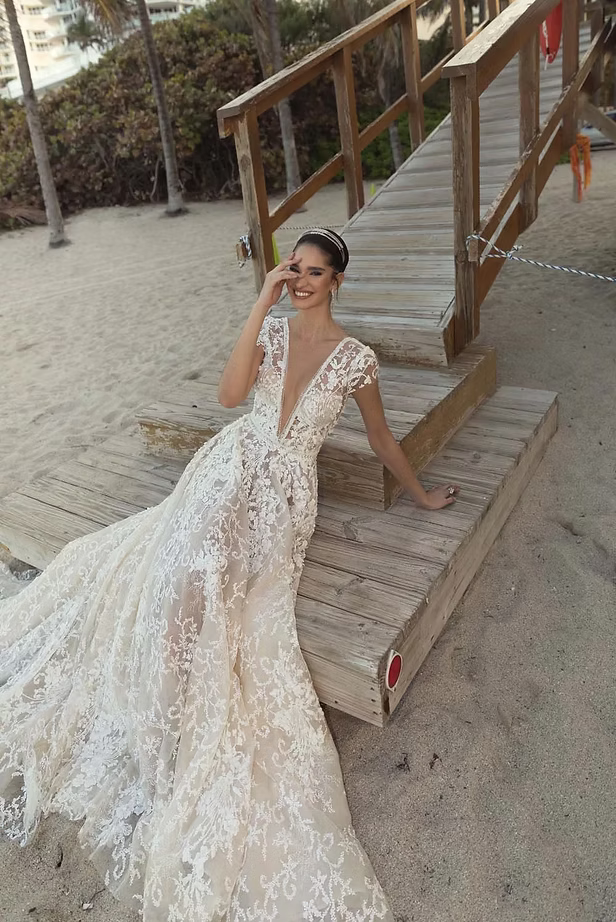 Woman in a lace wedding dress smiles, posed on a wooden beach walkway.