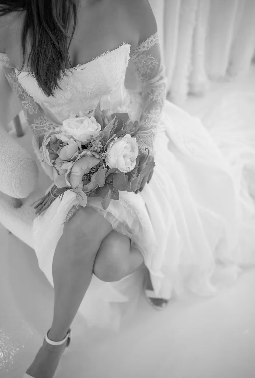 Bride in a white off-shoulder dress, holding a bouquet, sitting with legs crossed.