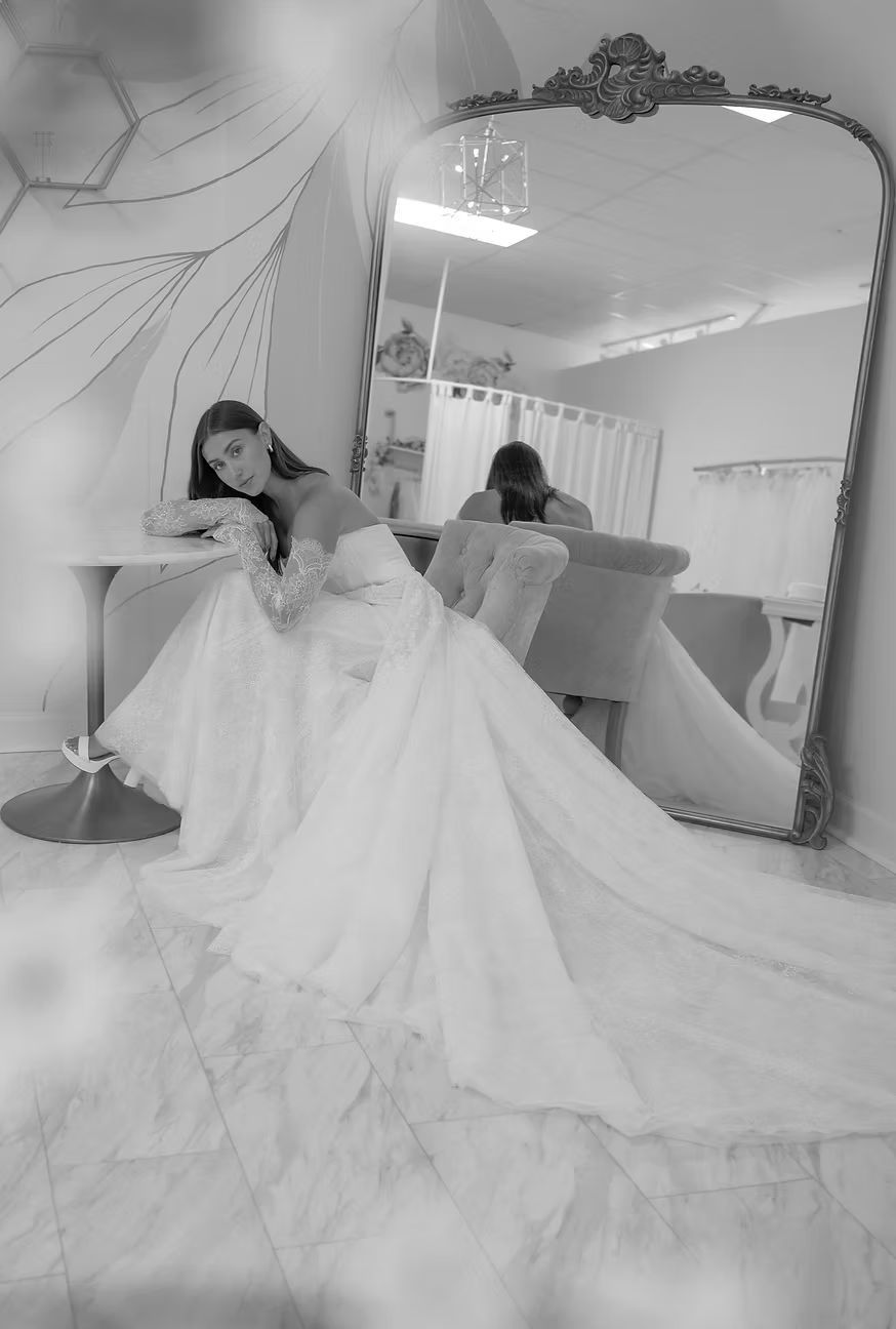 Bride in white gown leans on table, posing next to ornate mirror in a bridal shop.