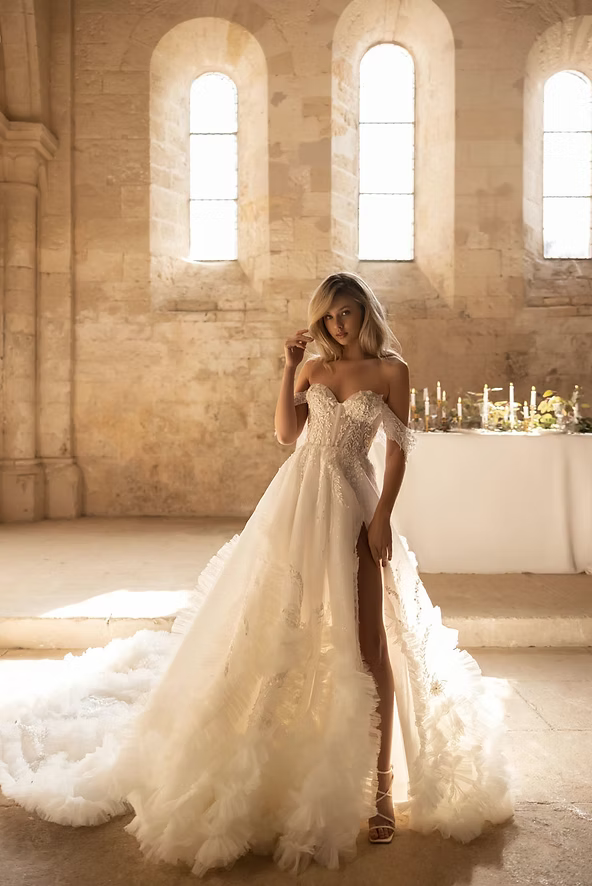 Woman in off-the-shoulder wedding dress with slit, standing in a sunlit stone building.