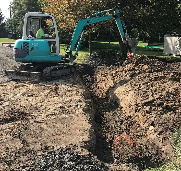 Small teal excavator digging a trench in dirt, person in the cab. Outdoors.