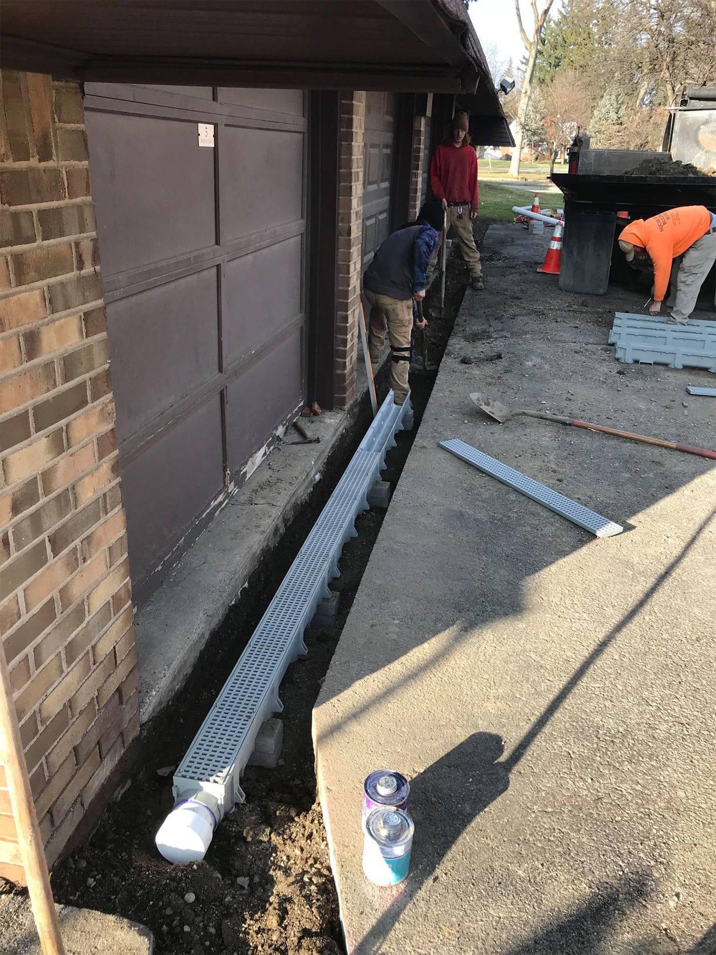 Workers installing a trench drain along a building. Brown garage door and brick exterior.