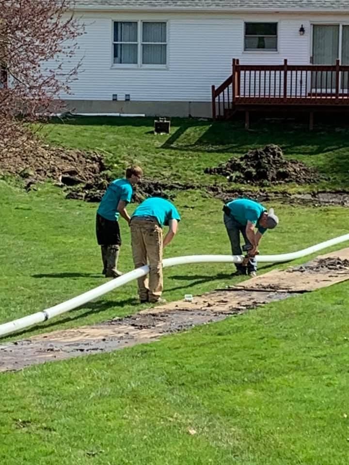 Three people in teal shirts install a white pipe on a lawn, near a house with a deck.